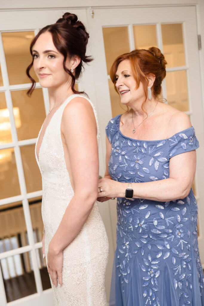 Bride smiles softly as her mother, dressed in a beaded periwinkle gown, lovingly fastens the back of her wedding dress in a bright room before the ceremony.