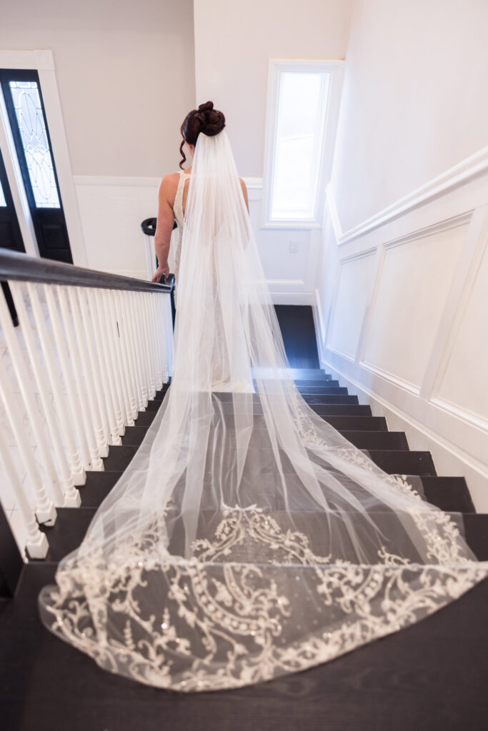 Bride walking down staircase with cathedral-length embroidered veil trailing behind, showcasing intricate lace details and elegant bridal styling at Hiland Park Country Club.