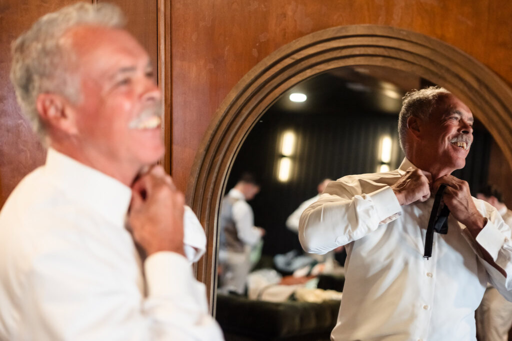 Father of the bride smiling as he adjusts his tie in the mirror, getting ready for the wedding day at Hiland Park Country Club surrounded by family and groomsmen.