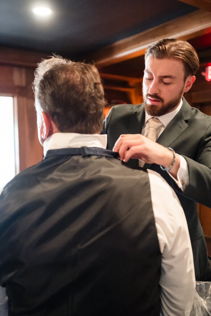 Groomsman helps adjust another man's tie in a warm, wood-accented room at Hiland Park Country Club, capturing a quiet moment of camaraderie during wedding day preparations.