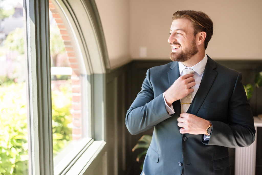 Groom smiling as he adjusts his tie near a sunlit window at Hiland Park Country Club, dressed in a tailored suit with a gold tie clip and leather watch.