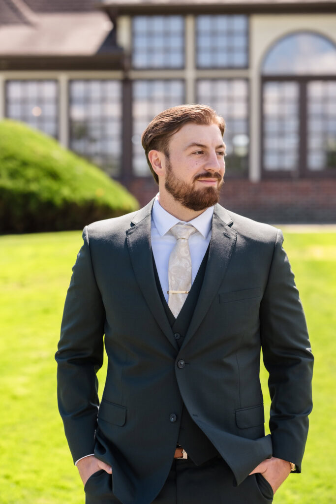 Groom standing confidently on the lawn at Hiland Park Country Club, dressed in a classic dark suit with a paisley tie and gold tie clip, with the venue’s elegant architecture in the background.