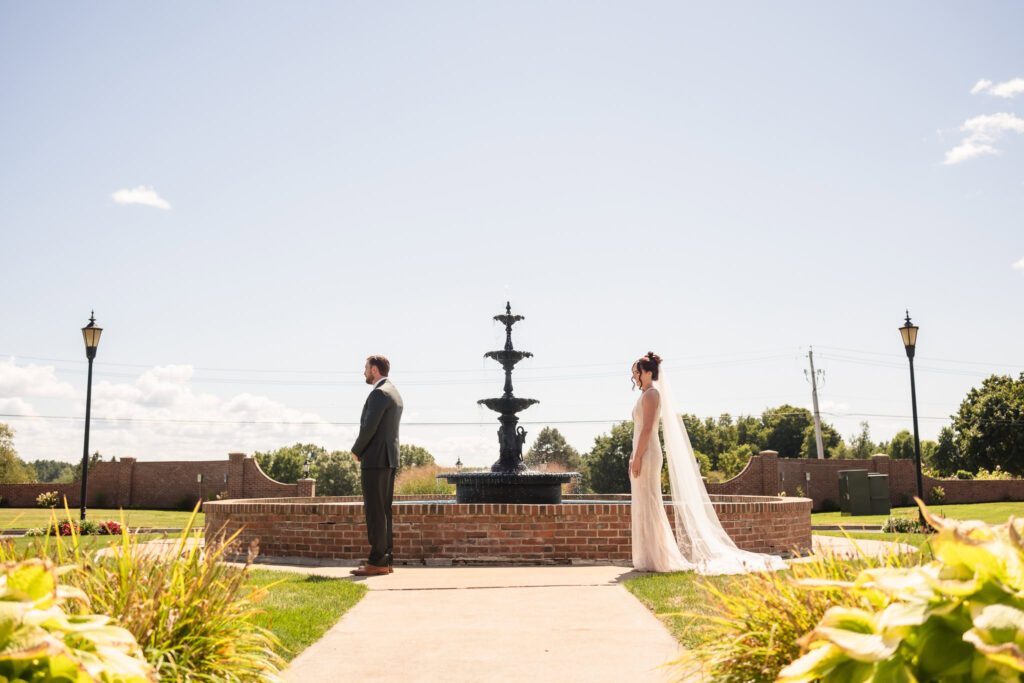 Bride and groom standing back-to-back in anticipation for their first look near the fountain at Hiland Park Country Club, framed by sunny skies and manicured gardens.