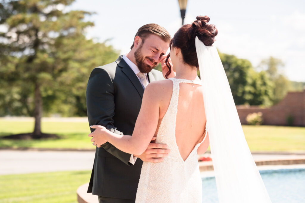 Groom overcome with emotion during first look, holding his bride close and wiping away tears in a heartfelt moment by the fountain at Hiland Park Country Club.