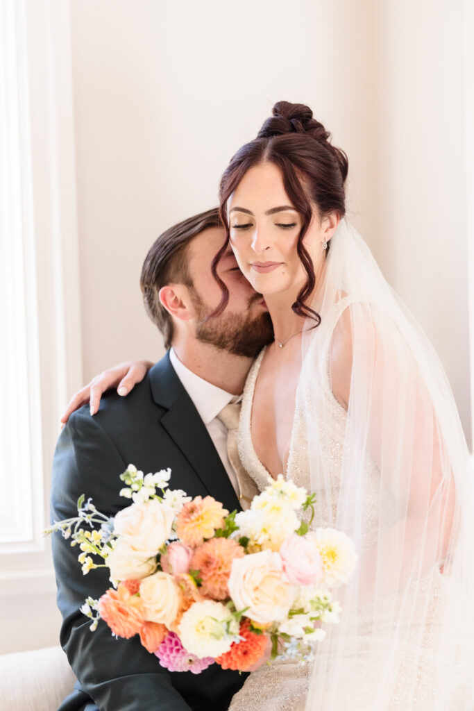Groom kisses his bride’s shoulder in a quiet, intimate moment by the window with the bride holding a lush bouquet of pastel and coral blooms.