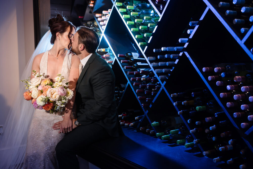Bride and groom share a romantic kiss in the wine cellar at Hiland Park Country Club, surrounded by dramatic lighting and rows of wine bottles, with the bride holding a vibrant floral bouquet.