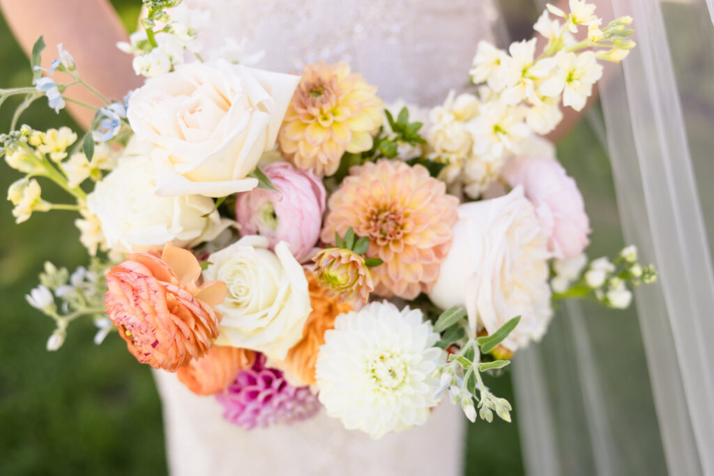 Close-up of bride’s romantic summer bouquet featuring white roses, peach ranunculus, blush dahlias, and soft pastel blooms, designed by Oopsydaisy Flower Farm for a Hiland Park wedding.