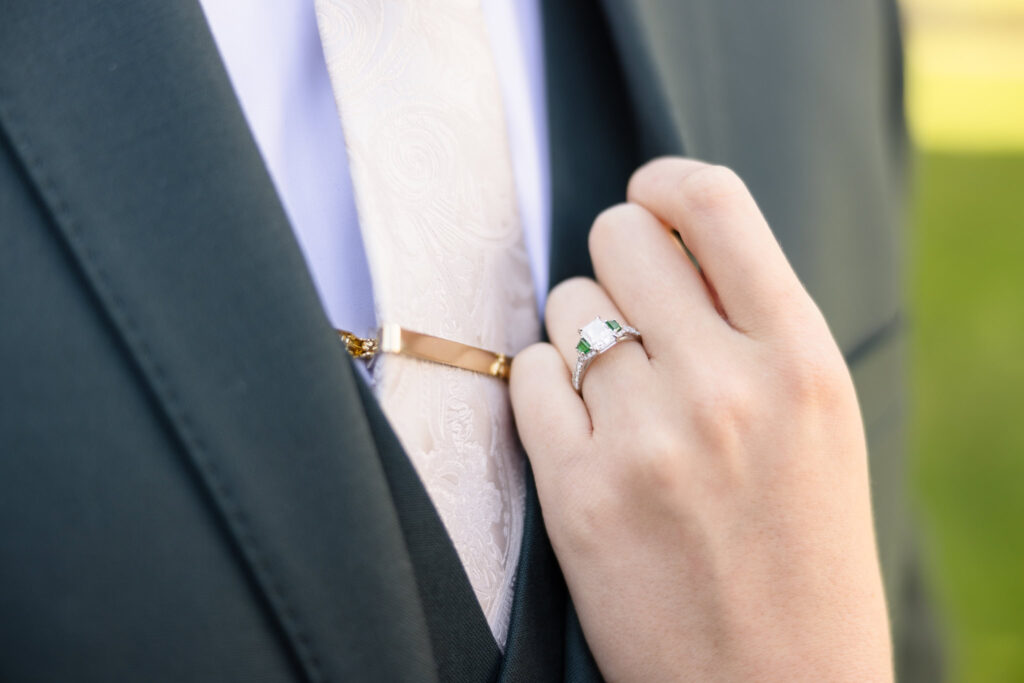 Close-up of bride’s hand resting on groom’s suit, showcasing her emerald-accented engagement ring and his ivory paisley tie with gold tie clip at their Hiland Park Country Club wedding.