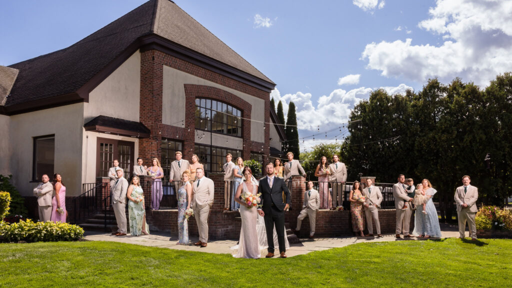 Full wedding party portrait outside Hiland Park Country Club, with the bride and groom centered among bridesmaids in pastel gowns and groomsmen in tan suits, set against the venue’s classic architecture and string lights.