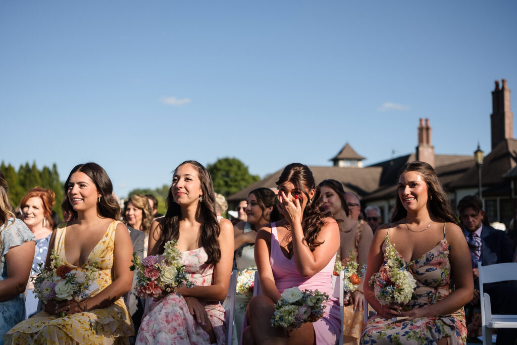 Bridesmaids seated during outdoor ceremony at Hiland Park Country Club, holding pastel floral bouquets and reacting with emotion and joy as they watch the couple exchange vows.