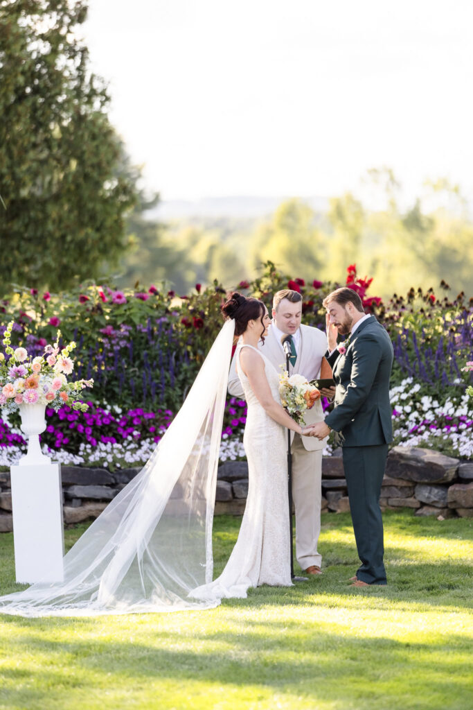Bride and groom exchange vows during a heartfelt outdoor ceremony at Hiland Park Country Club, surrounded by vibrant summer florals and officiated by the groom’s brother.