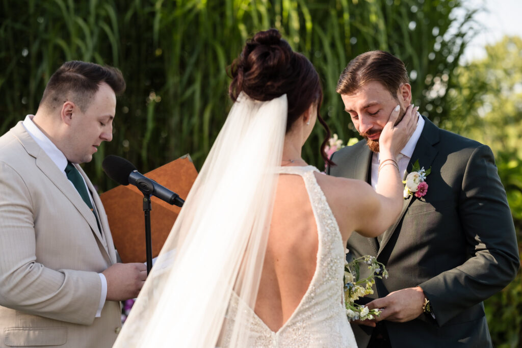 Bride gently touches the groom’s face as he tears up during their emotional wedding vows, with the officiant reading beside them at Hiland Park Country Club.