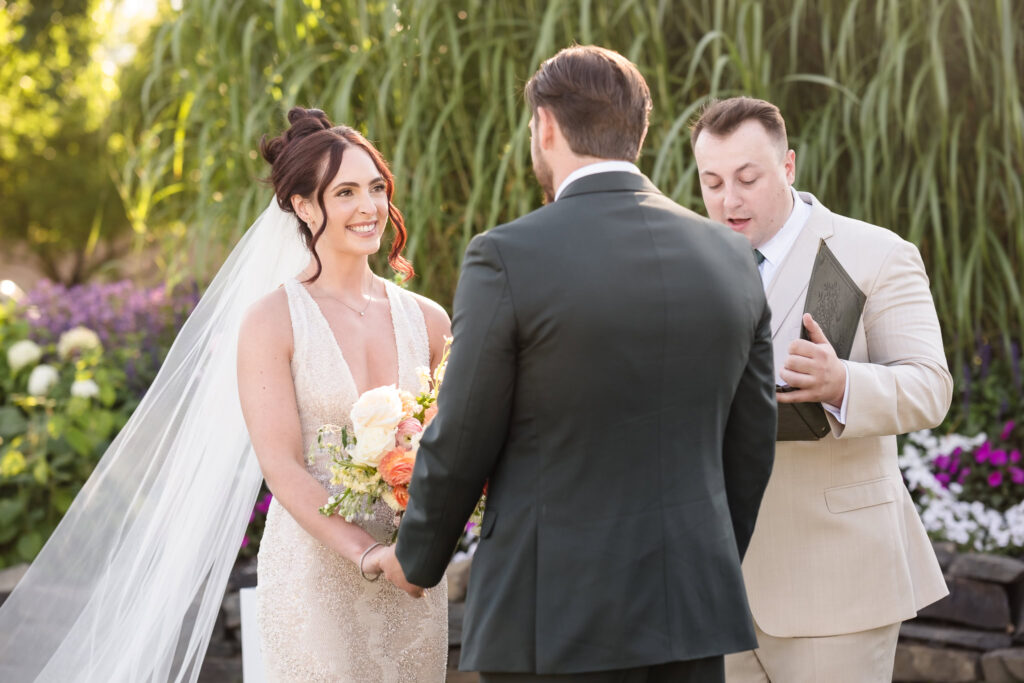 Bride smiles radiantly at her groom during their outdoor ceremony at Hiland Park Country Club, holding hands and bouquet as the officiant reads their vows.
