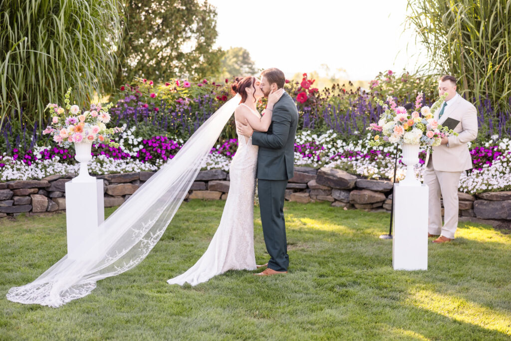 Bride and groom share their first kiss as husband and wife during a vibrant outdoor ceremony at Hiland Park Country Club, surrounded by summer florals and lush garden views.