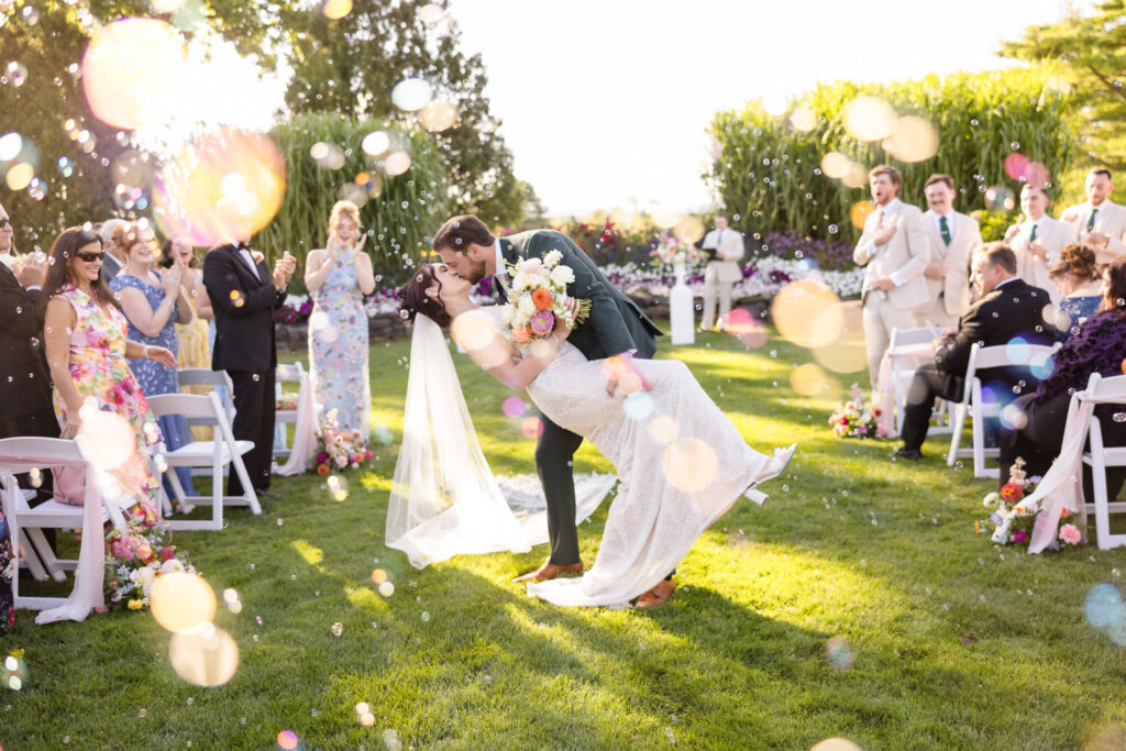 Bride and groom share a joyful dip kiss as they exit their ceremony at Hiland Park Country Club, surrounded by bubbles, cheers, and golden afternoon light.