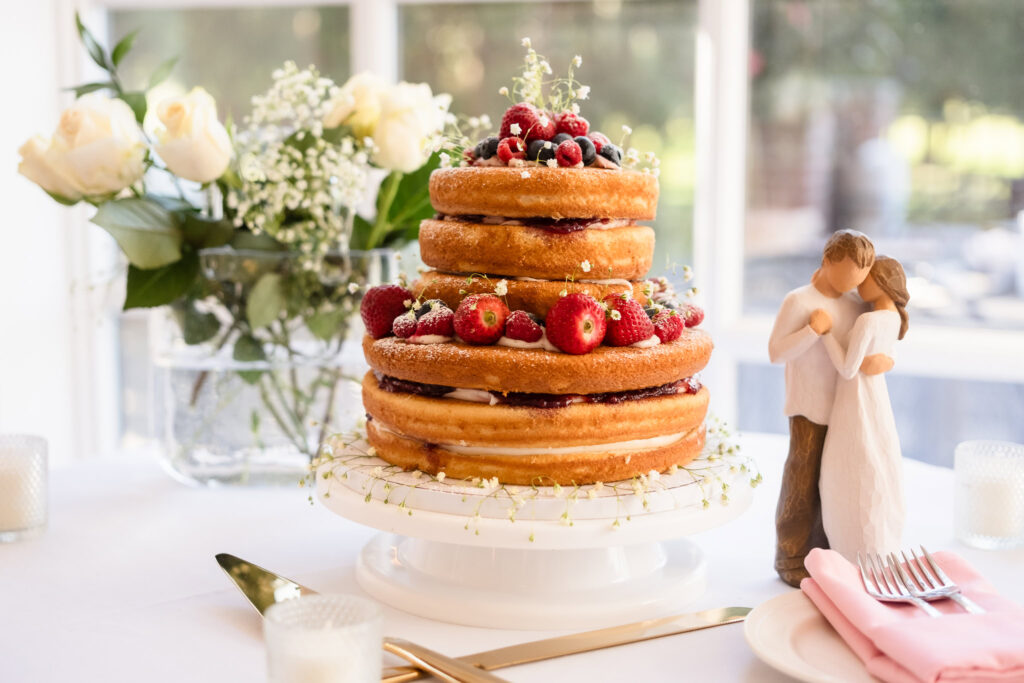 Rustic naked wedding cake adorned with fresh berries and baby's breath, displayed beside a romantic figurine and white roses.