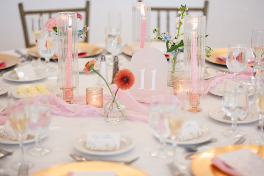 Elegant wedding reception table, featuring blush pink taper candles, gold chargers, delicate floral accents, and a soft chiffon table runner with Table 11 signage.