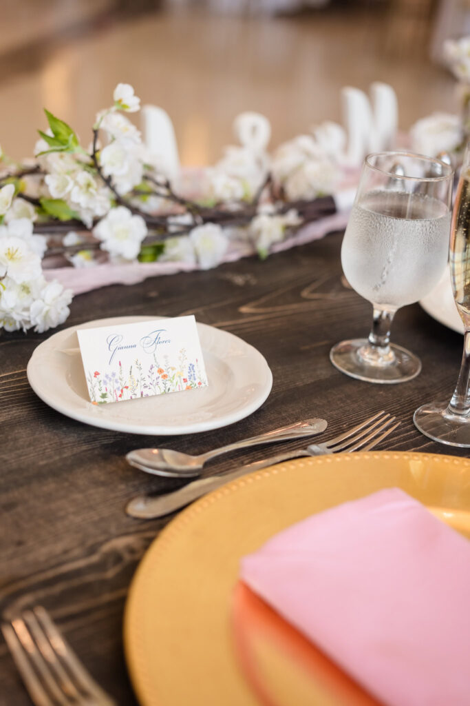 Close-up of elegant wedding place setting, featuring a gold charger, pink napkin, floral name card, and cherry blossom garland on a dark wood table.