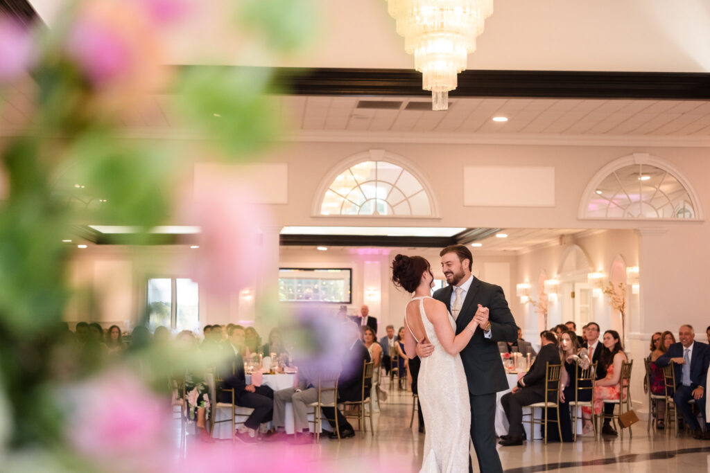 Bride and groom share their first dance at Hiland Park Country Club, smiling at one another as guests look on in a softly lit ballroom filled with floral details and golden accents.