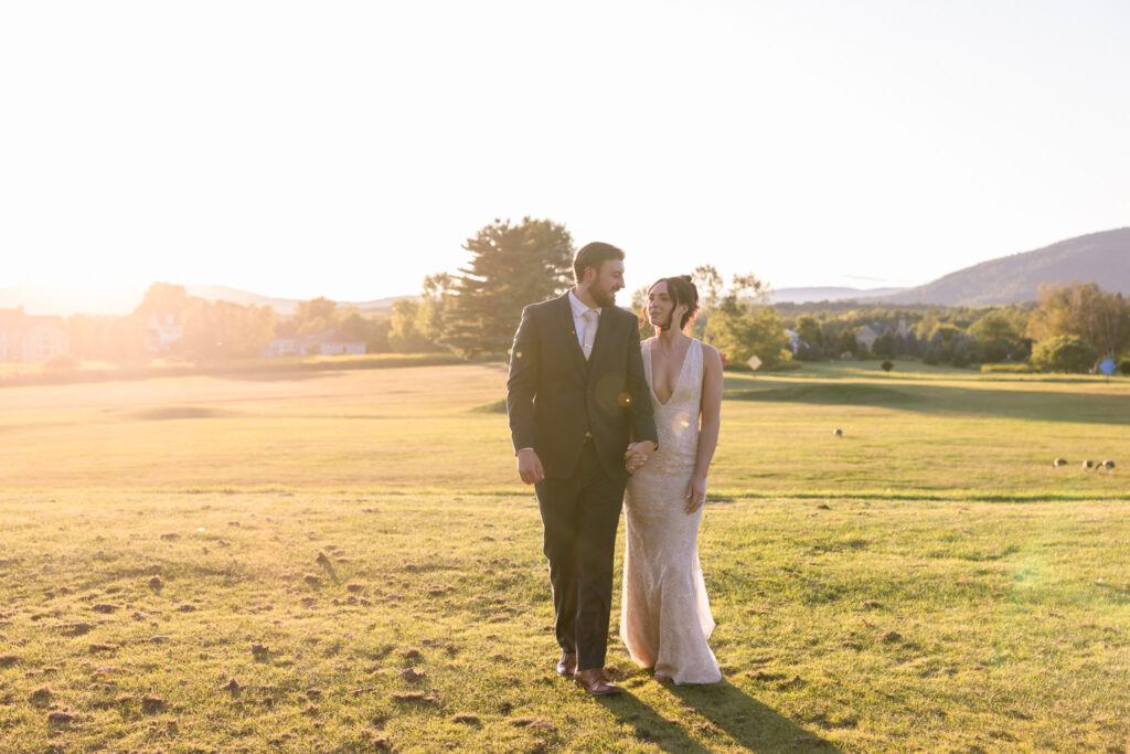 Bride and groom walk hand in hand across the sun-drenched golf course at Hiland Park Country Club during golden hour, with the Adirondack mountains in the background.