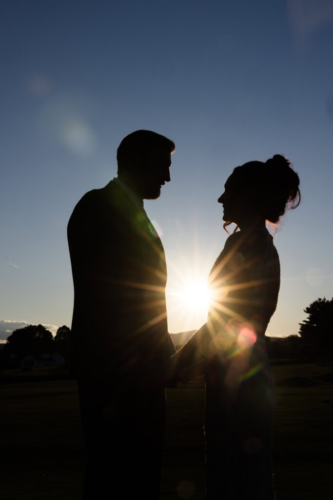 Silhouette of bride and groom holding hands at sunset on the golf course, with sun flare bursting between them in a romantic golden hour moment.