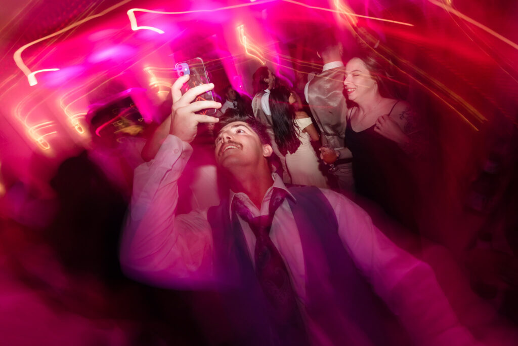 Wedding guest captures the moment on his phone amid vibrant pink lighting and high-energy dancing during the reception at Hiland Park Country Club.