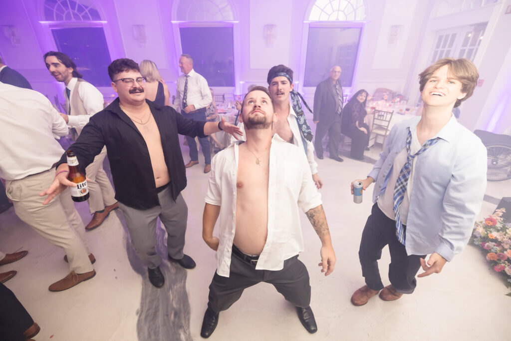 Group of wedding guests with open shirts and drinks in hand dancing wildly under purple lighting during a fun-filled reception at Hiland Park Country Club.