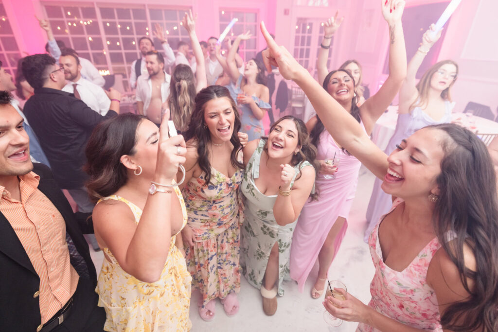 Guests in colorful floral dresses dance and sing with hands in the air under pink lights during a lively wedding reception at Hiland Park Country Club.