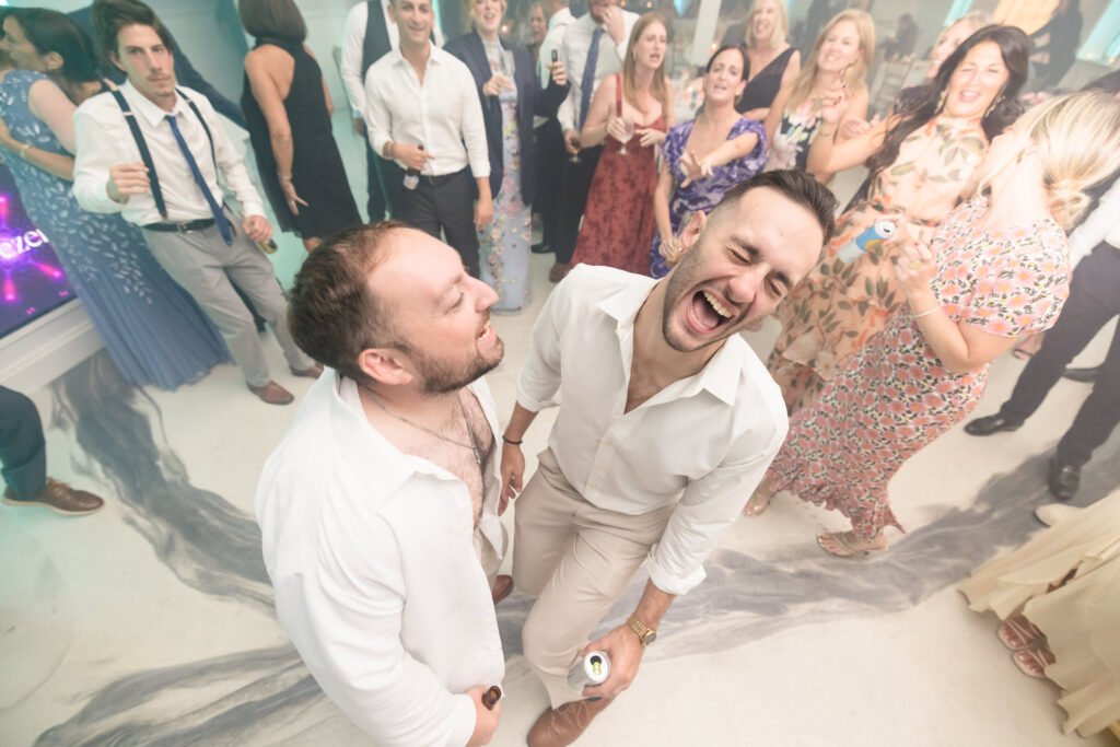 Two groomsmen burst into laughter on the dance floor surrounded by guests having a blast during the wedding reception.