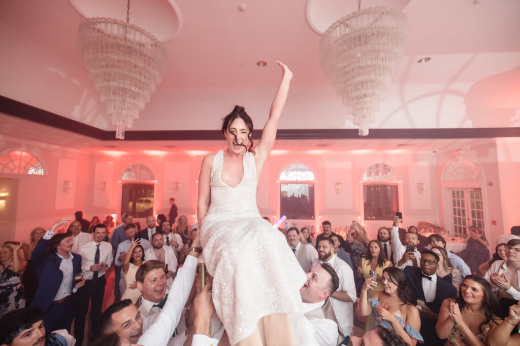Bride is lifted into the air on a chair during the Hora dance, beaming with excitement as wedding guests cheer and celebrate at Hiland Park Country Club reception.