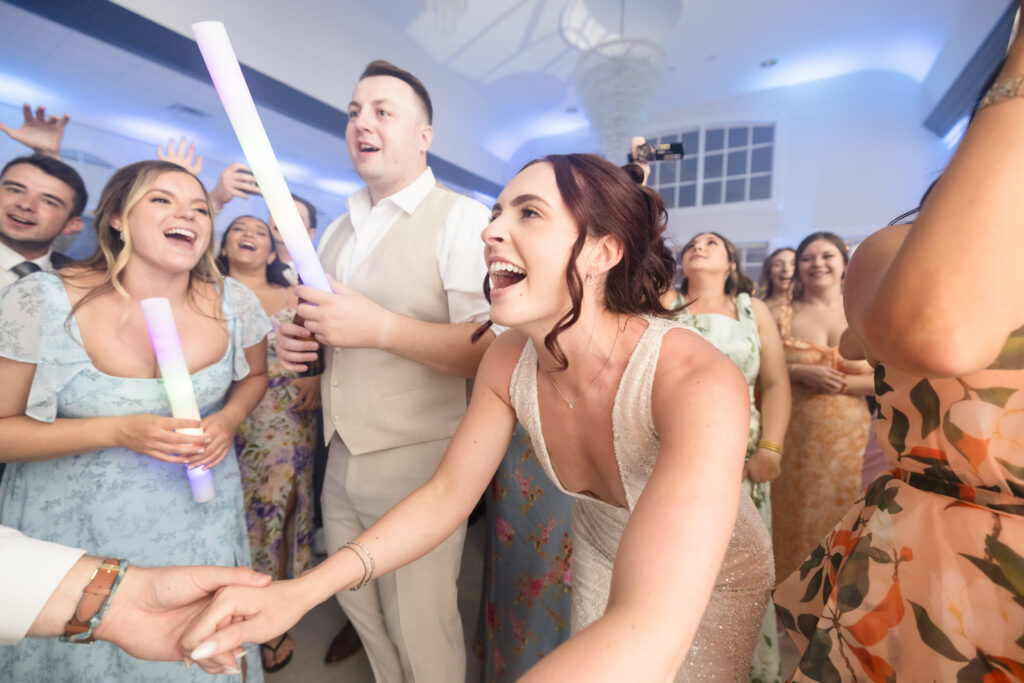 Bride laughs and reaches out joyfully on the dance floor surrounded by guests holding glowing light sticks during an energetic wedding reception at Hiland Park Country Club.