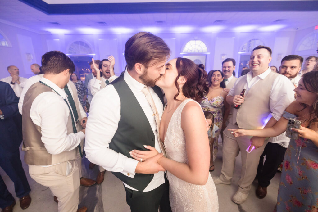 Bride and groom share a kiss on the dance floor surrounded by cheering guests during a joyful wedding reception.