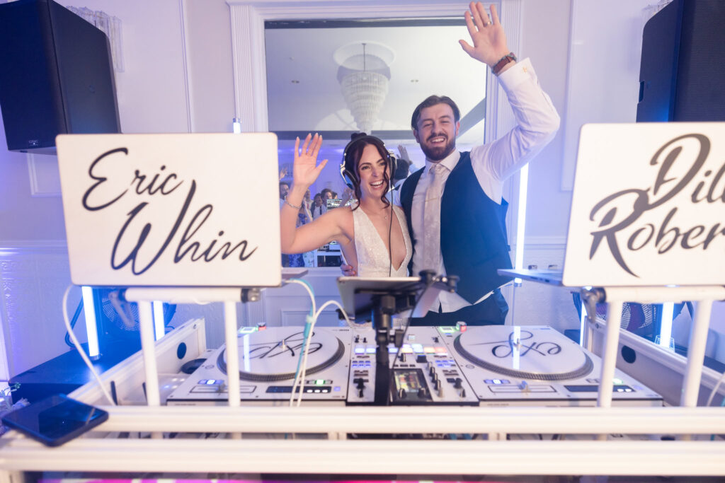 Bride and groom smile and wave behind the DJ booth at their wedding reception, celebrating with DJ Eric Whin and Dylan Roberts.