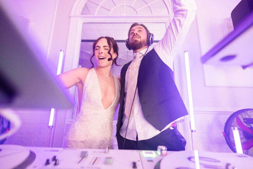 Bride and groom sing and dance behind the DJ booth, wearing headphones and glowing with excitement during their epic reception party.