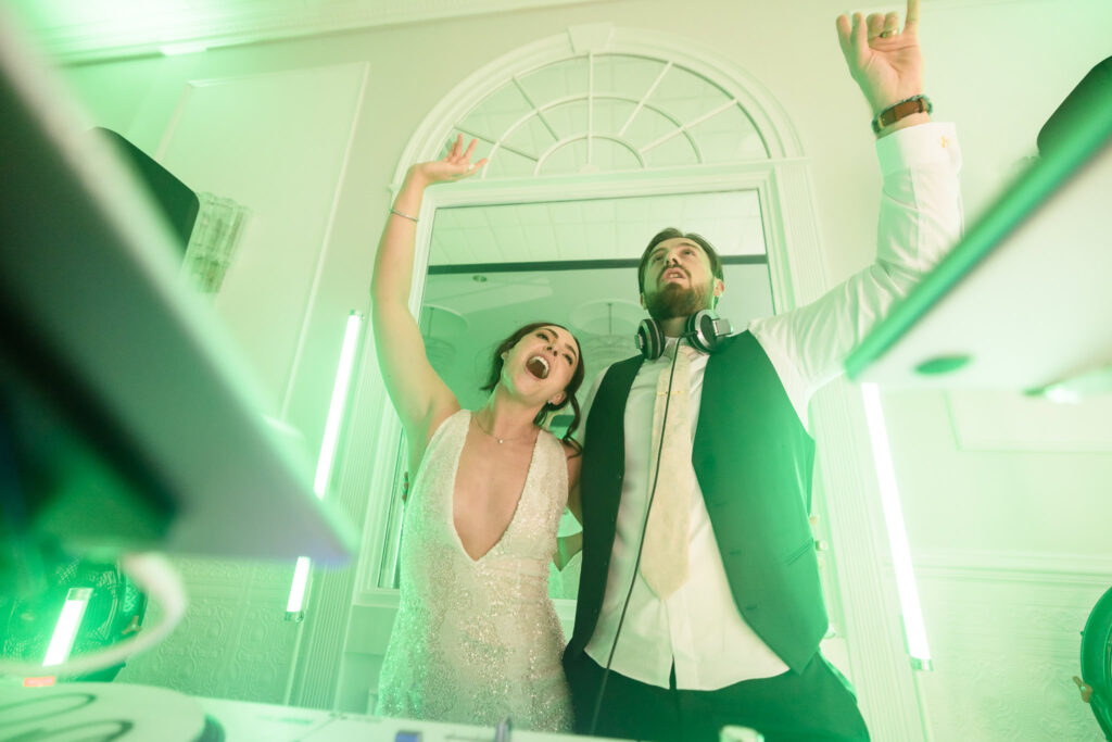 Bride and groom throw their hands up and sing with excitement behind the DJ booth, lit by vibrant green lights during their unforgettable wedding reception.