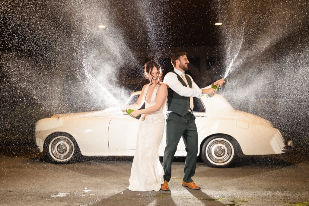 Bride and groom celebrate with a dramatic champagne spray in front of a vintage white car at night, marking the end of their wedding day at Hiland Park Country Club with laughter and flair.