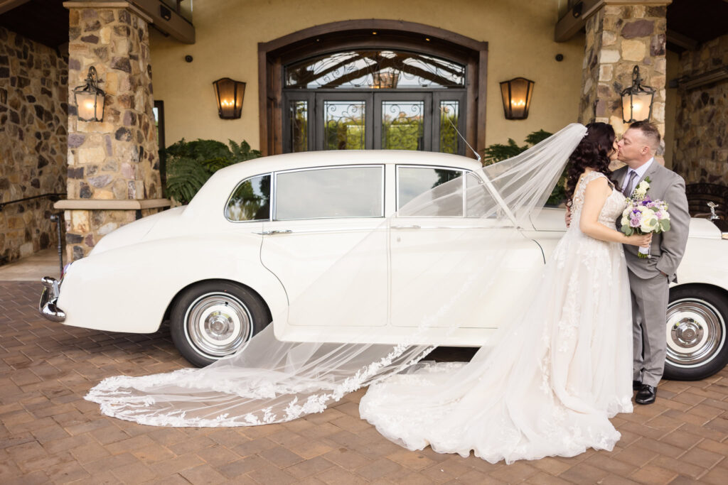 Bride and groom share a kiss in front of a vintage white Rolls-Royce at Larkfield Manor, with the bride’s long lace veil draped elegantly across the brick driveway.