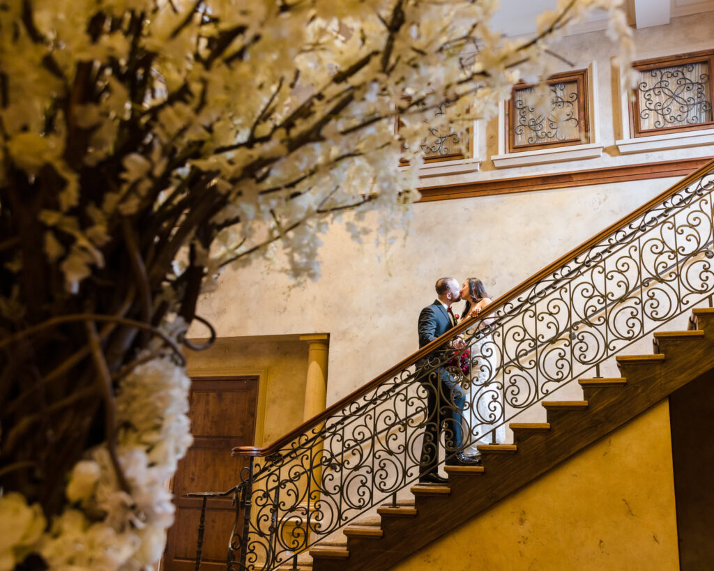Bride and groom share a romantic kiss on the grand staircase at Larkfield Manor, showcasing the venue’s elegant European-inspired architecture in East Northport, Long Island.