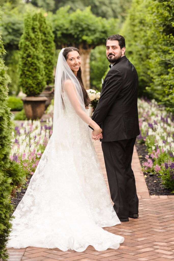 Bride and groom holding hands and smiling in the flower-lined brick walkway at Larkfield Manor, surrounded by lush gardens and Tuscan-inspired architecture.