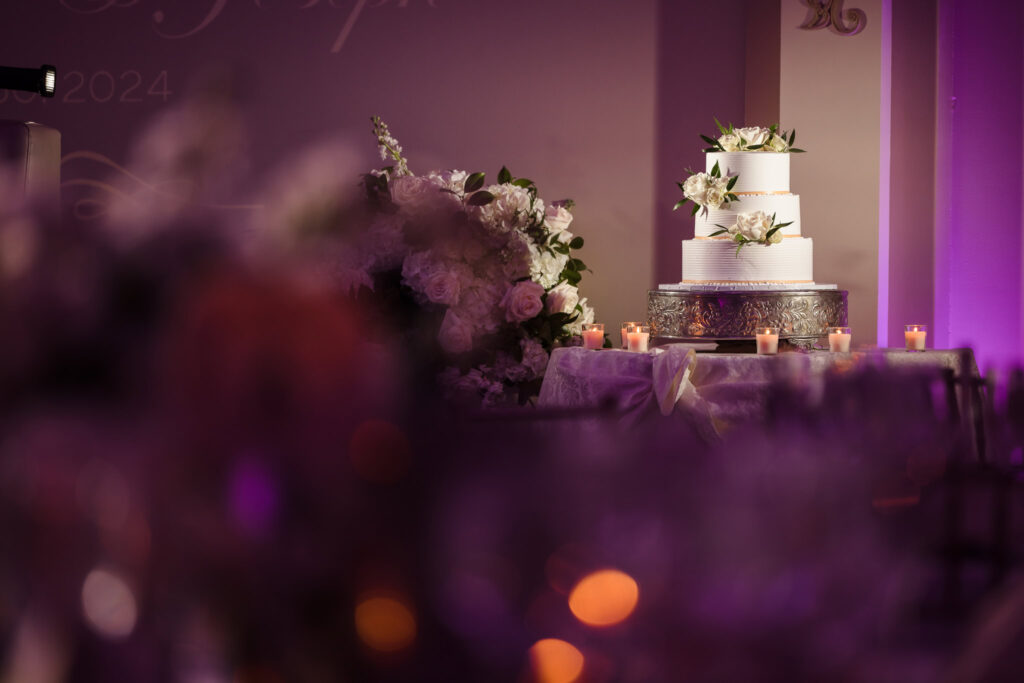 Elegant three-tier wedding cake with white floral accents displayed on a silver stand, surrounded by candles and romantic purple lighting.