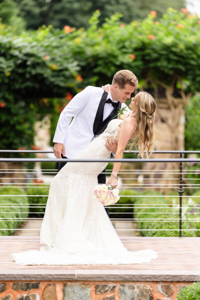 Groom in white tuxedo dips bride for a romantic kiss during garden portraits at Larkfield Manor wedding on Long Island.