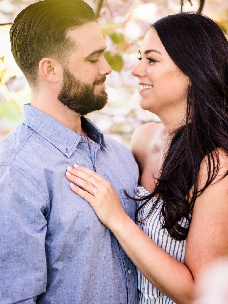 Romantic close-up engagement photo of a couple smiling at each other under spring blossoms, showcasing the bride’s engagement ring.