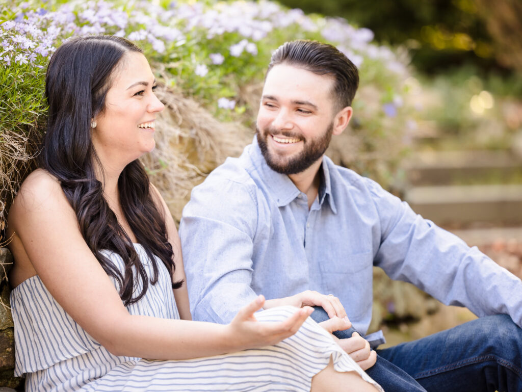 Candid engagement photo of a couple laughing together while sitting near blooming flowers