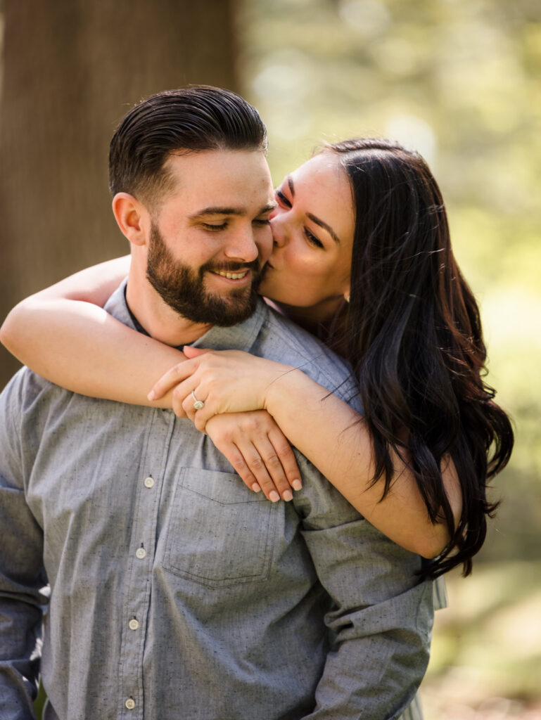 Playful engagement photo of a couple embracing in the gardens, with the woman kissing her partner’s cheek and showing her engagement ring.