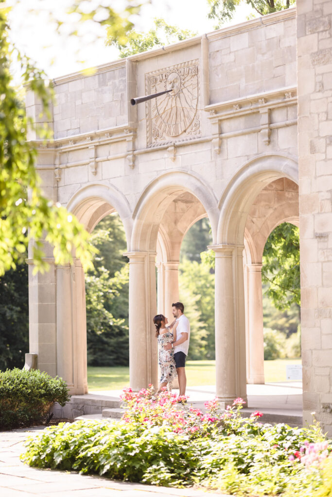 Elegant engagement photo of a couple standing beneath the grand stone arches at Planting Fields Arboretum, surrounded by summer greenery and blooming flowers.