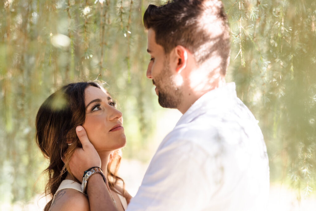 Tender engagement moment of a couple standing under soft sunlight and greenery at Planting Fields Arboretum in Oyster Bay, New York.
