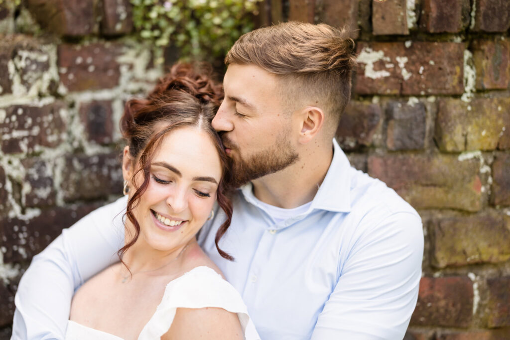 Romantic engagement portrait of a couple embracing and sharing a kiss on the forehead in front of a rustic brick wall