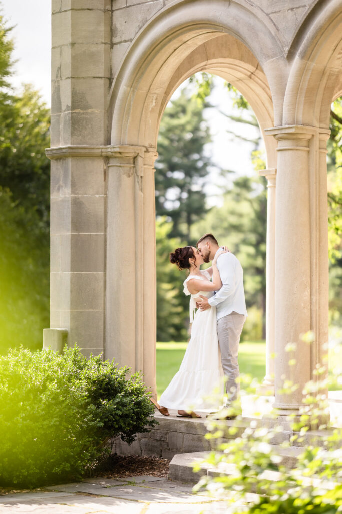 Romantic engagement photo of a couple sharing a kiss beneath the stone arches of Coe Hall Mansion at Planting Fields Arboretum in Oyster Bay, New York.