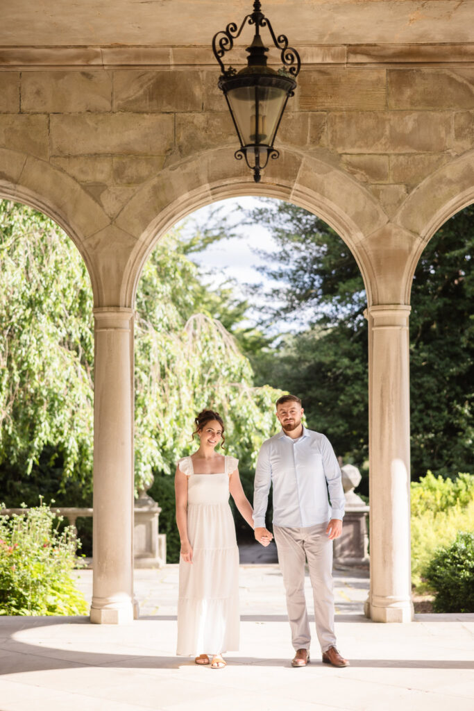 Elegant engagement photo of a couple holding hands beneath the stone arches of Coe Hall Mansion at Planting Fields Arboretum in Oyster Bay, New York.