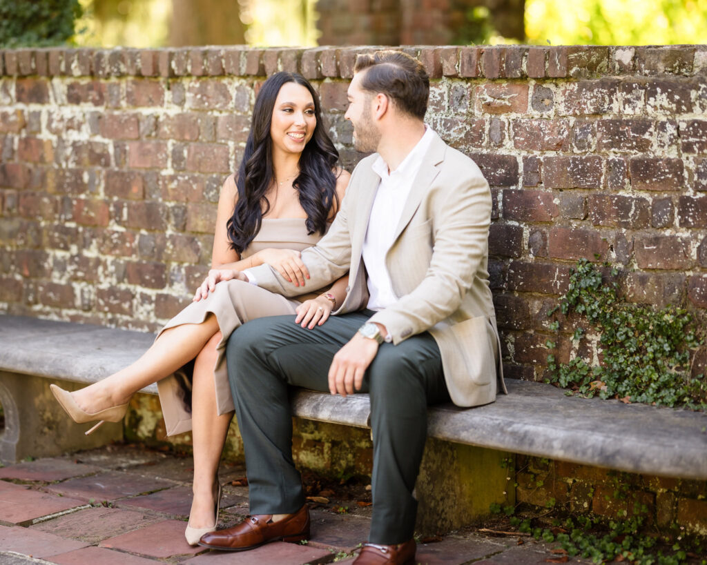 Elegant engagement photo of a couple sitting on a stone bench against a brick wall at Planting Fields Arboretum, sharing a warm and intimate moment in the garden.